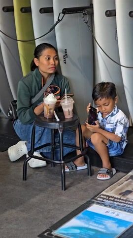 A woman and a young boy are sitting on the ground next to a row of surfboards. The woman is sitting on a low stool, wearing jeans, a green jacket, and white sneakers. She has her hair pulled back. A handbag is slung over her shoulder. On the stool are two plastic cups of iced drinks, one topped with whipped cream. The boy is sitting on the ground looking at a smartphone. He is wearing a blue-and-white patterned shirt, blue shorts, and sandals. The surfboards are leaning against a wall.  A printed sign can be seen at the bottom right of the frame. The setting appears to be inside a shop or cafe.