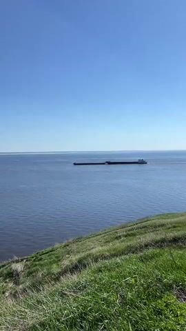 The video shows a wide river with a long cargo ship on it under a clear blue sky. The camera zooms out to reveal a grassy hill in the foreground, and a man in a white tank top, shorts, and a baseball cap. He walks into the frame and takes a photo of the river with his phone. The sun is shining brightly, causing a glare on the water.
