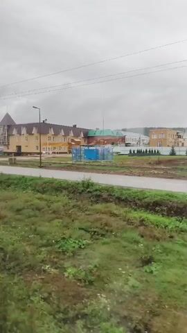 The video shows a view from a moving vehicle, likely a train or bus, looking out a window. In the foreground, a dark vertical pole is visible, partially obscuring the view. Beyond the pole is a landscape with green grass and vegetation. In the middle ground, there is a road and some railway tracks. On the other side of the road and tracks, there are several buildings. The most prominent building is a light-colored, multi-story structure with a distinctive pointed roof and large windows. Other buildings, including what appear to be industrial or commercial structures, are visible further in the distance. The sky is overcast and grey. The video is shaky, indicating movement.