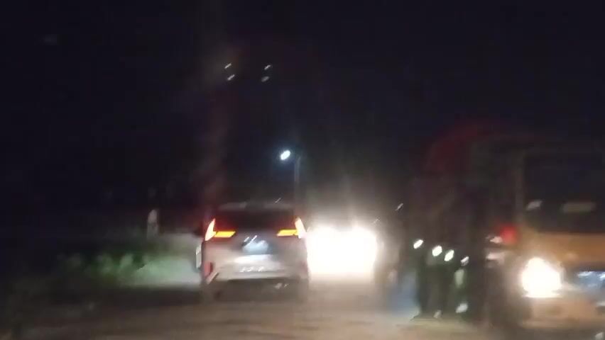 This video shows a view from inside a vehicle driving at night on a road.  The road appears to be dirt or gravel.  In front of the vehicle, a white SUV with its taillights illuminated is visible.  Further ahead, oncoming headlights of at least two vehicles are bright and blurry. To the right of the white SUV and slightly ahead is a large truck with its side marker lights visible.  There are streetlights and other light sources in the distance. The sky is dark.