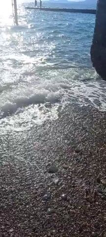 This video captures a serene beach scene at Gwandra, Abkhazia. The camera is positioned low, near the pebbled shore, as gentle waves lap at the beach. The sunlight glints off the water's surface, creating a shimmering effect.

In the distance, a pier extends into the sea, with several indistinct figures standing on it, possibly enjoying the view or fishing. The ocean appears calm and inviting, with clear blue water. The weather seems pleasant, likely daytime given the bright sunlight. The overall mood is tranquil and peaceful, evoking a sense of relaxation. The video focuses on the natural beauty of the coastline and the gentle rhythm of the waves.