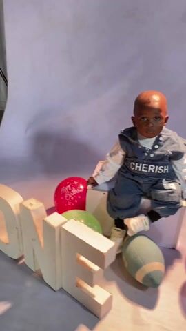 A baby sits on a white box in a studio setting, surrounded by props for a first birthday photoshoot. The baby is wearing a light denim outfit with "CHERISH" printed on the front and white sneakers. Next to the baby are large, white block letters spelling "ONE," along with several balloons in various colors (red, green, purple, yellow) printed with "Happy Birthday." A green football also lies on the floor. The backdrop is a light purple gradient. The baby is looking down.