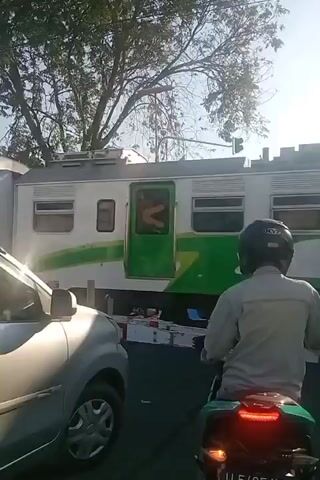 A daytime video captures a train passing through a level crossing. A silver car is stopped on the left side of the frame, partially obscuring a motorcycle rider in a light-colored shirt and dark helmet. The motorcycle is stopped behind the car, facing the train tracks. To the right of the first motorcycle is another motorcycle with a rider wearing a dark shirt and dark helmet. The train car visible is white with a green stripe. Trees and buildings are visible in the background. The sun is shining brightly, creating lens flare.