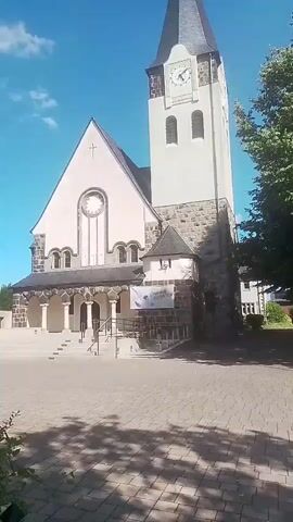 The video shows a church with a tall bell tower. The church has a large arched window above the entrance. There is a banner hanging on the side of the church. The building to the left of the church appears to be a community hall or school, with solar panels on the roof. The area in front of the church is paved with cobblestones. The sky is blue with some clouds. The video is shot from ground level, moving slightly to the left.