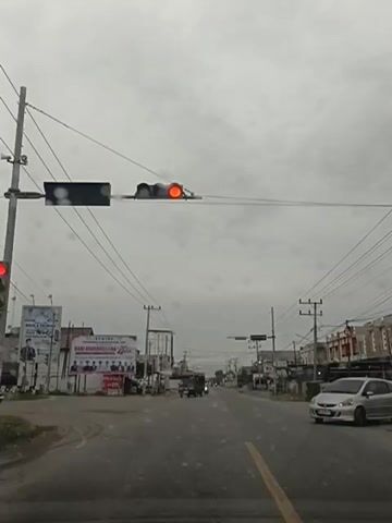 The video shows a frontal view of a street scene on an overcast day. The camera is positioned inside a vehicle, as indicated by the perspective. A traffic light is visible overhead, displaying a red and yellow light, indicating a caution or upcoming stop. Power lines crisscross the sky and are supported by poles on either side of the street. On the left side of the street, there is a billboard with images of people. Further down the road, there are several businesses and buildings. A few vehicles, including cars and motorcycles, are present on the road. The road surface appears to be asphalt, with a yellow line dividing the lanes. There are some raindrops visible on the windshield, suggesting it may be raining or has recently rained. The overall atmosphere is subdued due to the cloudy sky.