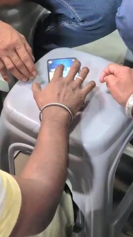 The video shows a close-up view of a smartphone placed on a grey plastic stool. Three hands are visible, interacting with the phone or the stool. The phone screen displays a colorful interface, possibly a game or an app with multiple icons. One hand, with a silver bracelet and a ring on one finger, is actively touching the screen. Another hand, with a silver bracelet, is resting on the stool near the phone. A third hand, with a blue denim-clad leg in the background, is reaching towards the phone or the stool. The environment appears to be indoors, with a shallow depth of field that blurs the background.