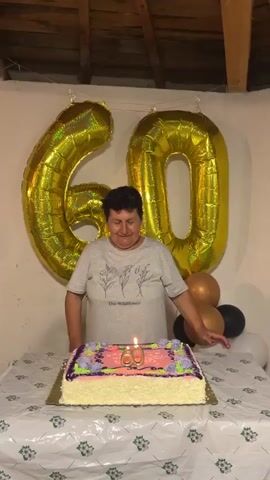 In a celebratory scene, a woman stands behind a table adorned with a birthday cake and festive decorations. The cake, rectangular in shape, is frosted with pink and white icing, and bears the number "60" in the center, complete with a lit candle. Behind the woman, two large gold foil balloons spelling out "60" are hung, and a cluster of gold, white, and black balloons are positioned to the side. The woman, wearing a gray t-shirt with a floral design, beams at the camera and moves her hands in a celebratory manner. The setting appears to be indoors, with exposed wooden beams forming the ceiling.