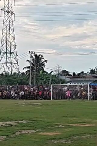 A soccer game is underway, with a crowd gathered behind a goal and a lone player in pink, presumably the goalkeeper, positioned in front of the net. A soccer ball is visible on the green field in the foreground, indicating the game is in progress. In the background, a large electrical tower and some buildings suggest an urban or semi-urban setting. The sky is cloudy, hinting at a potential change in weather.