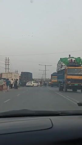 The video, shot from the interior of a vehicle, captures a busy intersection in an urban setting. Multiple trucks, some heavily laden with goods, dominate the scene, interspersed with cars and motorcycles. Several people are walking across the street, adding to the congestion. Overhead, utility poles and streetlights line the road. The overall tone suggests a bustling, potentially chaotic traffic environment. The sky appears overcast, indicating possible weather conditions.