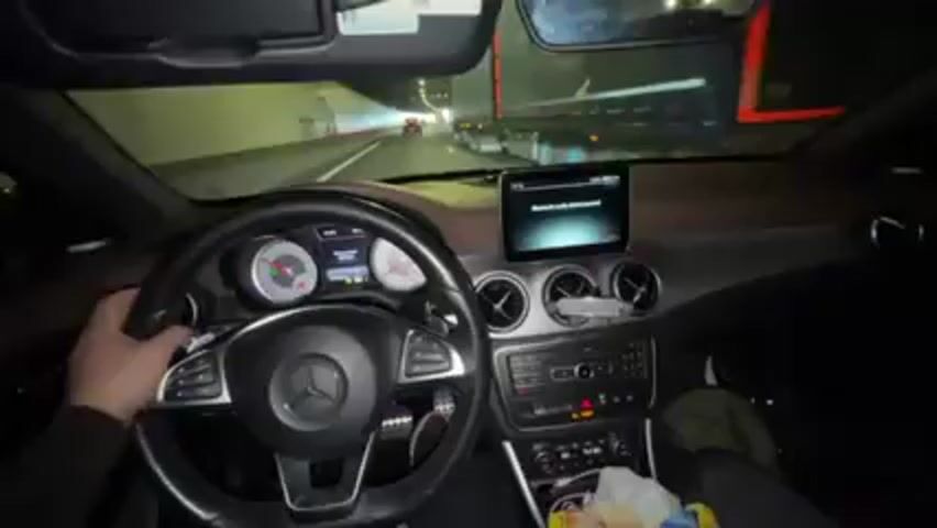 The interior of a moving Mercedes-Benz CLA-Class is shown from the driver's perspective. The driver, whose hands are visible on the steering wheel, is navigating through a brightly lit tunnel on a multi-lane highway. Other vehicles, including a large truck, are visible ahead and to the right. The car's dashboard features a central display screen, air vents, and various buttons. A bag of snacks sits in the passenger seat.
