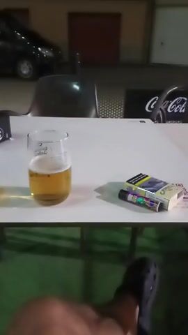 The video shows a table with a glass of beer, a pack of cigarettes, and a lighter. A person's legs and feet are visible in the foreground. The background is blurred but appears to be an outdoor seating area with a car and a Coca-Cola sign. The lighting suggests it is evening or night. The video is approximately four seconds long and appears to be a short clip or a looped segment.