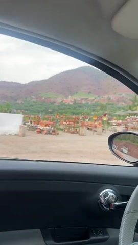 The video, filmed from inside a moving car, captures a series of roadside establishments against a backdrop of red-brown mountains and green vegetation in Taljarft, Morocco, on an overcast day.

The scene initially shows outdoor stalls displaying numerous terracotta pots, vases, and other decorative pottery items on wooden stands. Wooden tables and chairs are set under thatched umbrellas, suggesting outdoor dining or resting areas. In the background, a village with scattered buildings and a prominent light-colored structure is visible on the mountainside. The car's side mirror reflects the road behind, showing a truck.

As the car continues, a building with walls made of woven material and a red-tiled roof comes into view, alongside a large, circular woven object leaning against it. Further along, a wooden kiosk-like structure with a red roof is seen, with a motorcycle parked nearby. People are visible sitting at tables under another thatched umbrella.

The landscape consistently features rolling, reddish-brown hills and mountains, interspersed with green patches. Towards the end, a decorative heart-shaped archway, seemingly constructed from small, multicolored pieces, stands in the background. The side mirror intermittently shows the road and other passing vehicles.