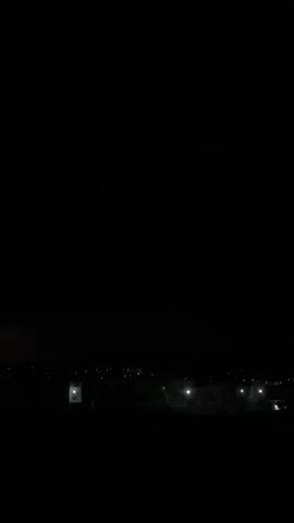 The video shows a night scene of a town or city from a distance. In the foreground, there is complete darkness. Further away, one can make out the silhouettes of buildings and the sparse lights of streetlamps and houses. The scene is still and there's no discernible movement.