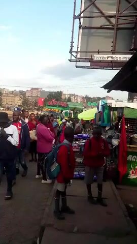 The video shows a street scene with a group of people. In the foreground, two young boys in red school uniforms with backpacks are standing on a raised concrete area next to a market stall. Behind them, a crowd of adults, some wearing red shirts, are walking and gathered. There are market stalls with goods displayed, including clothing. In the background, buildings and a billboard are visible. The sky is cloudy.