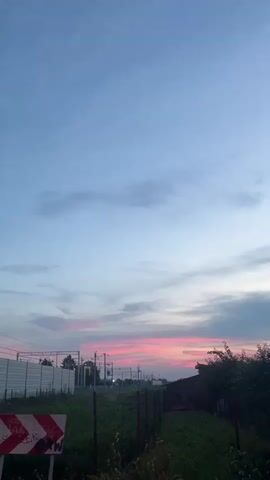 This is a vertical, low-angle shot of a rural or suburban landscape at dusk. The sky dominates the frame, transitioning from a pale blue at the top to hues of pink and orange near the horizon, indicating sunset. Wispy clouds are scattered across the sky, some catching the sunset's glow.

In the middle ground, a fence runs horizontally, separating a patch of green grass from a more distant area. Beyond the fence, there are visible signs of human infrastructure. A white noise barrier wall is prominent on the left, with train tracks and overhead power lines stretching into the distance. Several poles and what appear to be small buildings or sheds are scattered throughout this area. On the right side of the frame, a dark-colored building with a sloped roof is partially visible, nestled amongst some trees. The overall setting suggests a location near a train line, possibly on the outskirts of a town or village.

In the foreground, a red and white chevron sign is visible on the left, indicating a warning or directional marker. The sign has some graffiti on it. The grass in the immediate foreground is overgrown.

The time of day is clearly dusk, with the soft, warm colors of the sunset painting the sky. The weather appears calm, with no strong winds suggested by the foliage or clouds. The emotions evoked are generally peaceful and contemplative, due to the serene sky and the quiet atmosphere implied by the scene. No people are visible in the image. The location is identified as Ożarów Mazowiecki, Poland.