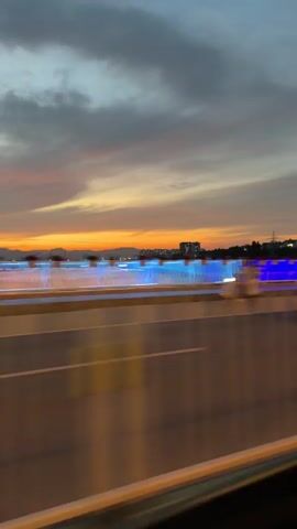 The video captures a fleeting view from a moving vehicle, possibly a car or bus, on a bridge during what appears to be sunset or dusk. The sky is painted with hues of orange, yellow, and gray, with clouds streaking across it. In the background, a silhouetted cityscape with buildings and lights can be seen against the horizon. The bridge itself is illuminated with vibrant blue lights along its railing, creating a striking contrast with the warm tones of the sky. There are some blurred figures, possibly pedestrians or other vehicles, visible on the bridge. In the foreground, the road surface and the vehicle's interior are visible, adding to the sense of motion and perspective. A prominent feature in the video is a tall lamppost with red lanterns, adding a touch of cultural aesthetic to the scene. The overall impression is one of a beautiful and dynamic urban landscape at twilight, experienced from a moving perspective.