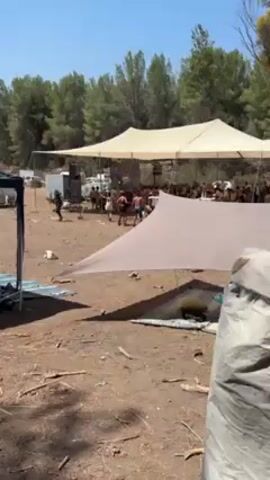 The video shows a daytime outdoor party under a large, light-colored tent. A crowd of people, most wearing minimal clothing, are gathered together, dancing and socializing. Several smaller shade structures are visible around the main tent. In the background, a lush forest provides a natural backdrop, while vehicles and equipment suggest a temporary setup. The ground is dry and littered with debris. A large sound system is set up behind the crowd.