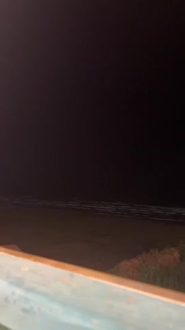 A dark, wide shot of the ocean at night. The horizon line is faintly visible, marked by a lighter strip of water where waves are breaking. In the foreground, the edge of a weathered, turquoise-painted wooden structure runs horizontally across the bottom of the frame. The scene is dimly lit, suggesting it is either very early morning, late evening, or heavily overcast.