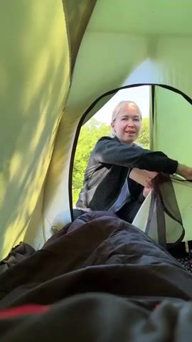 The video is filmed from inside a light green camping tent, looking outwards. A blonde woman, appearing to be in her 30s or 40s, is seen from the chest up, positioned just outside the tent's main opening. She wears a dark, patterned long-sleeved shirt and has a cheerful smile on her face.

At the beginning, she is opening the tent's zippered entrance. Bright daylight illuminates the scene, casting soft shadows on the tent fabric and suggesting it is morning or late morning. Lush green trees and foliage are visible in the background, indicating an outdoor, natural setting, likely a forest or campsite in Shirokaya Schel, Russia.

After opening the main tent flap, the woman reaches in to close the inner mesh screen, briefly obscuring her face. She then reappears, still smiling, holding two pink cups. Inside the tent, a dark sleeping bag or blanket is visible, along with a textured sleeping pad. A tag with the word "NORWAY" can be briefly seen on an item inside the tent. Her actions suggest she is either greeting someone inside the tent or bringing items in, possibly for a morning beverage during a camping trip.