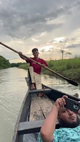 A video captured from a moving boat shows a young man with short, dark hair, wearing a maroon t-shirt with the word "Ferrari" in white on the front and light-colored pants with a black stripe down the side. He is sitting on the edge of the boat, holding a long wooden pole with his right hand. His left foot is on the wooden floor of the boat, and his right foot is bent. The boat is on a body of water, and on the right side of the frame, a person's head and shoulders are visible, holding a black smartphone. The background shows lush green vegetation and a cloudy sky with the sun partially visible. The boat appears to be moving forward.
