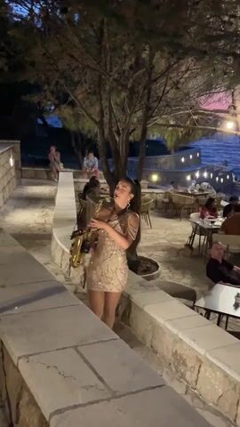 A young woman in a sequined dress plays a saxophone in an outdoor restaurant setting at dusk. She stands on a stone pathway, surrounded by trees and tables with diners. The restaurant appears to be near a body of water, possibly the sea, with lights reflecting on the surface. The overall atmosphere is relaxed and elegant.