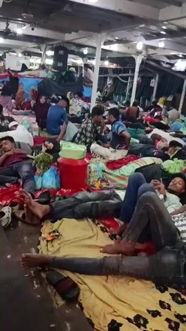 The video shows a crowded indoor space, likely the inside of a ferry or large boat, filled with people resting and sitting on blankets and mats spread across the floor. Many people are lying down, some asleep, while others are sitting and talking. There are personal belongings scattered around, including bags, containers, and shoes. The space appears to be brightly lit with overhead fluorescent lights. The overall atmosphere seems crowded and potentially cramped, with people maximizing the available space for rest and relaxation during the journey.