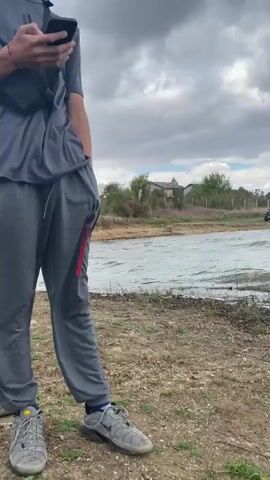 A video captures a cloudy day at a body of water, possibly a lake. In the distance, a dark-colored car is parked near a fence. The foreground shows choppy water lapping at a dirt and grass shoreline. Two individuals are visible fishing from the shore. The first individual, wearing a camouflage shirt and shorts, stands further away from the camera, casting a fishing line. The second individual, closer to the camera and wearing a black shirt and shorts, is also fishing. A fishing rod lies on the ground in the lower right corner of the frame, next to a backpack and a white container. The sky is filled with dramatic, heavy clouds.
