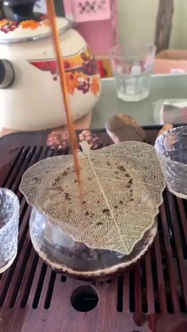 A close-up video captures a tea ceremony. A dark liquid is being poured from a thin stream into a small, round, dark ceramic bowl that sits on a wooden tea tray. A delicate, translucent leaf-shaped object rests on top of the bowl, acting as a lid. The liquid creates bubbles as it fills the bowl, and the stream of liquid continues to pour. In the background, a white teapot with a fall-themed pattern is visible, along with several small glass cups. The overall setting appears to be indoors, with soft lighting.