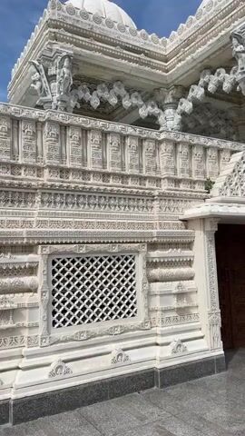 A video shows the exterior of a white marble temple, possibly Hindu, with intricate carvings and sculptures. The camera pans across the facade, revealing architectural details such as domes, columns, windows with decorative grilles, and relief carvings of figures. The sky is visible in the background, with some clouds. There are no people visible in the video. The overall impression is one of grandeur and detailed craftsmanship.