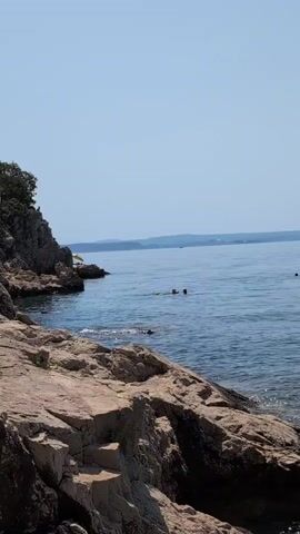 The video shows a rocky coastline with clear blue water and a clear sky. In the distance, there are several people swimming in the ocean. The foreground is dominated by large, rough-hewn rocks, suggesting a natural, undeveloped beach area. The water appears calm with gentle ripples.