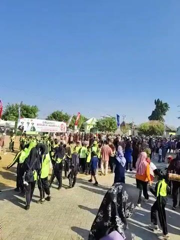 A video displays a large gathering of people outdoors on a sunny day. The crowd appears diverse, with individuals of various ages and genders, many wearing casual attire. Some people are wearing matching outfits, such as bright green vests or red jackets, suggesting they may be part of a group or event. In the background, there are buildings and trees, and a flagpole with a flag is visible. The overall atmosphere seems to be one of community engagement or a public event.