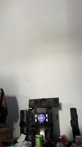 This media file is a short video clip showing a black stereo system with multiple speakers and a central unit displaying "BLUE" in red text. A circular blue light is illuminated on the central unit. The stereo system is positioned against a plain, light-colored wall, possibly in a home or room setting. There are no people visible in the clip. The lighting appears to be artificial, and the overall scene is static, suggesting the media file is a simple recording of the object. There are no discernible location cues other than the plain wall and the stereo system itself. The time of day and weather are not determinable. The mood is neutral and observational.