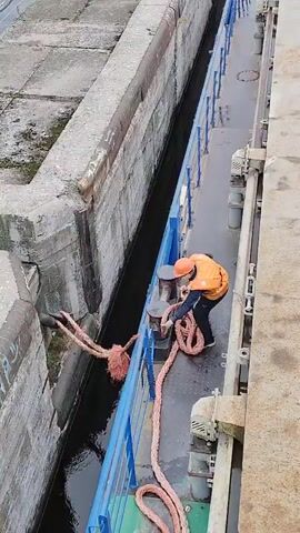 A person wearing an orange hard hat and a reflective vest is on a boat, holding onto a thick rope. The rope extends from the boat to a dock or a lock system. The person is pulling the rope, maneuvering it around a metal post on the deck of the boat. The water in the lock is dark and choppy. Concrete structures surround the lock. The scene appears to be in a shipyard or a port.