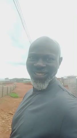The video is a close-up, selfie-style recording of a Black man with a grey beard. He is wearing a black t-shirt. The man is outdoors, with a dirt road and a low wall made of cinder blocks visible behind him. The sky is overcast. The man is making exaggerated facial expressions and speaking, but his words are not discernible.