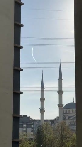 The view through an opening between buildings reveals a blue sky, power lines, the tops of two minarets and the dome of a mosque. A contrail arcs across the sky. A light breeze is possibly present as trees in the lower portion of the frame appear to sway.