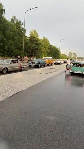 A collection of vintage cars are gathered on a wet asphalt road. The cars are various models and colors, including a green station wagon, a dark blue sedan, and an orange Volkswagen van. Some people are visible near the cars, with one person walking away from the green station wagon. The environment appears to be outdoors with trees lining the side of the road and a cloudy sky. The road is wet, suggesting it may have recently rained. The cars appear to be participating in a car show or parade.