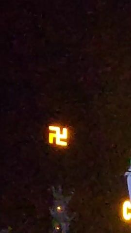 The video shows a dark outdoor scene with illuminated elements. A bright, orange swastika symbol is prominently displayed. Adjacent to it is a white statue of Buddha with the name "CHÙA LINH" in illuminated orange letters beside it.  A small decorative object, possibly a lit plant, sits at the bottom of the frame, closer to the swastika. The scene seems to be captured at night.