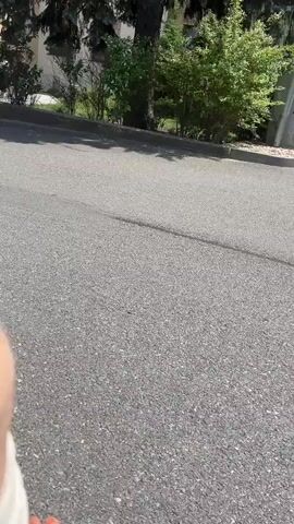 A first-person perspective video shows an asphalt road with tree shadows. In the background, there is a tan building with a blue "F" logo and windows, and a white car parked in front of it. A black car is also visible further down the road. The camera appears to be moving, possibly being pushed or pulled.