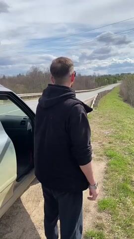 A man in a black hooded jacket and black pants gets out of a silver-colored car parked on the shoulder of a two-lane road. The road has a guardrail. The man walks a few steps, then jumps over the guardrail onto a grassy slope. In the background, there are trees and a lake. The sky is cloudy.