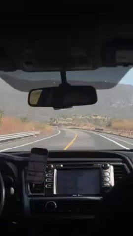 The view from inside a car shows a driver traveling down a two-lane road with a yellow centerline.  The landscape consists of low scrub vegetation and a small mountain range in the distance. A smartphone is mounted in a holder to the left of the car's infotainment system, which is not displaying information. The rearview mirror reflects a blurry image of a passenger. The car's interior is visible, including the dashboard and some of the front seats. The weather appears to be sunny and clear.
