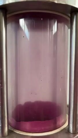 A close-up, vertical shot shows a clear cylindrical container, likely made of glass or plastic, encased by two silver metal poles on either side. Within the cylinder, a deep purple substance has settled at the bottom, forming a thick, irregular layer. Above this layer, a pale purple liquid fills most of the cylinder, with small dark particles dispersed throughout. The lighting suggests an indoor environment, possibly with natural light filtering from above, as evidenced by a faint reflection on the top rim of the container. There are no people visible in the frame, and the focus remains solely on the contents and container.