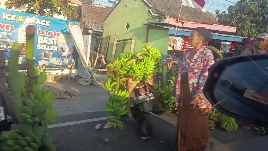 The video shows a street scene with several people and a large amount of bananas for sale. In the foreground, from the perspective of a car's side mirror, a woman wearing a floral shirt and brown skirt is seen standing next to a motorcycle laden with bunches of green bananas. To her right, a man in a brown jacket and red skirt is also standing near the bananas. Further back, other individuals appear to be involved in selling or arranging the bananas, with a makeshift stall visible behind them. The environment appears to be a roadside market with buildings and trees in the background. The overall atmosphere is that of a busy commercial area.