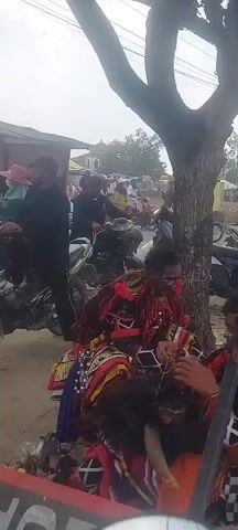 The video shows a street scene with many people and motorcycles. In the foreground, two individuals are dressed in colorful, elaborate costumes, possibly for a performance or cultural event. One person appears to be adjusting the costume of the other. Behind them, a crowd of people on motorcycles are gathered, some wearing helmets. A tree with large leaves is prominent in the mid-ground. In the background, there are buildings, banners, and more people. The overall atmosphere seems to be one of a public gathering or festival.
