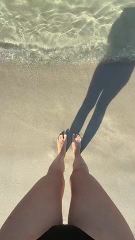 The video opens with a first-person perspective looking down at a person's bare feet standing at the edge of a sandy beach, with clear, shallow water lapping at their toes. The sunlight casts a long shadow of the person onto the sand and into the water. The water is translucent, revealing the sandy bottom. As the video progresses, the camera pans up and out, revealing a wider view of a beautiful bay.

The scene is a tropical or Mediterranean beach with calm, crystal-clear turquoise water. The sand is light in color and appears soft. In the distance, there are rocky outcrops and hills covered in vegetation. Several sailboats are visible anchored in the bay, suggesting a peaceful and leisurely atmosphere. The sky is a vibrant blue with a few wispy clouds, indicating clear, sunny weather. The time of day appears to be late morning or early afternoon, given the angle of the sun and the length of the shadows.

The activity is simple: a person is enjoying a moment at the beach, perhaps wading into the water or preparing to swim. The overall mood is one of relaxation, tranquility, and enjoyment of nature's beauty. The clarity of the water and the bright sunlight evoke a sense of warmth and serenity. No visible text is present in the media file. The location context of Es Camp De Mar, Spain, suggests a scenic coastal resort.