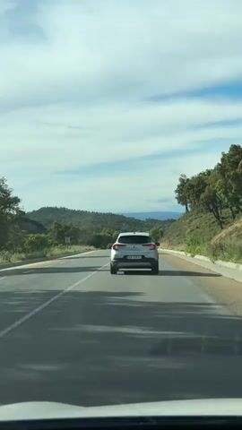 A car is driving on a two-lane highway. The sky is partly cloudy, and there are rolling hills with trees in the distance. Another car is driving in the opposite direction, and a silver vehicle is in front of the car recording the video.