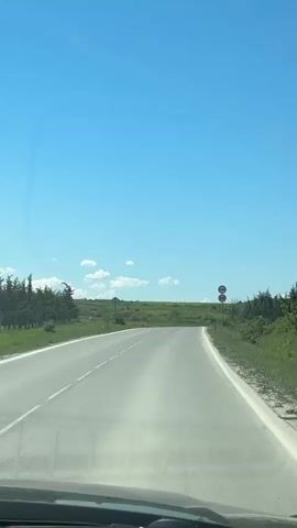 The video is shot from the dashboard of a moving vehicle driving on an empty asphalt road. The sky is a clear blue with a few wispy white clouds.  The road appears to be in a rural area with green fields and scattered trees and bushes on either side. There are two round traffic signs with red borders indicating "no overtaking" and a speed limit of "50" visible on the right side of the road.  The road is slightly uphill. The dashboard is visible at the bottom of the shot.  A snippet of music is audible.
