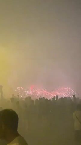 The video captures a crowd of people gathered at night, silhouetted against a hazy sky illuminated by fireworks. The fireworks are visible in the background, producing bursts of red and pink light and sparks. Many people in the crowd are holding up their phones, presumably to record the event. The atmosphere appears festive and celebratory. The lighting is dim and colored by the fireworks and some external artificial light source on the left side of the frame. The air is filled with smoke or haze, obscuring finer details of the environment and the faces of the people.