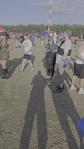 The video shows an outdoor event with a large crowd of people. In the background, there are several tents and stages, as well as a prominent windmill-shaped structure with smoke or mist emanating from it. People are scattered across a grassy field, some standing and observing, while others appear to be walking or interacting. The sky is clear and blue with scattered clouds, indicating daylight. The overall impression is of a festival or large gathering.