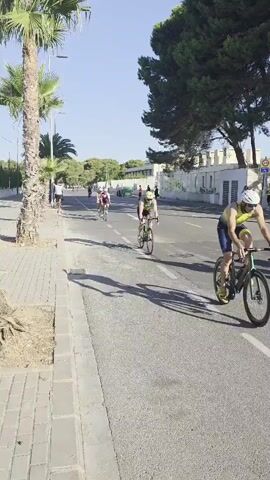 A low-angle shot captures a cycling race on a sunny day. In the foreground, a cyclist wearing a helmet and athletic wear rides a bicycle towards the camera, casting a shadow on the asphalt road. Palm trees line the left side of the road, their fronds reaching towards the clear blue sky. A sidewalk runs alongside the palm trees, paved with grey bricks. In the mid-ground, several other cyclists are visible, also wearing helmets and athletic gear, riding their bicycles in the same direction. The road is marked with white dashed lines, separating lanes. In the background, a low white wall with crenellations runs along the right side of the road, bordered by lush green trees. A few spectators can be seen in the distance, near the wall. The overall scene depicts an outdoor sporting event in a warm, possibly coastal, environment.