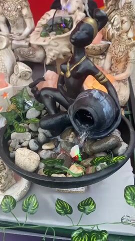 The video shows a table at an outdoor market displaying various decorative objects for sale. In the foreground is a small fountain featuring a black statue of a woman sitting and pouring water from a pot into a basin filled with smooth stones and artificial greenery.  Behind the fountain, there are several other decorative items including various Buddha statues, owl figurines, and salt lamps in white containers with star-shaped cutouts. In the background, a woman with dark hair is looking at the items on the table. The overall impression is of a collection of spiritual or New Age-themed decorative items for sale.
